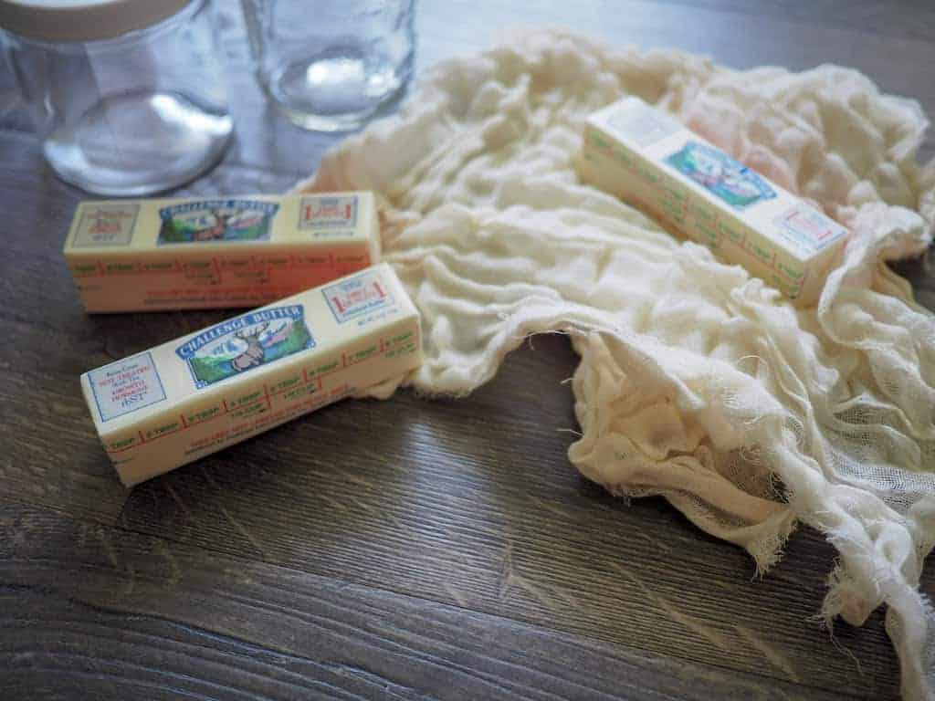 Two glass jars, three sticks of butter, and cheesecloth on a wooden surface.