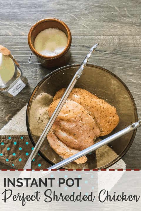 Raw chicken breasts in a brown glass bowl with metal tongs, salt, and herbes de provence on a gray wood floor.