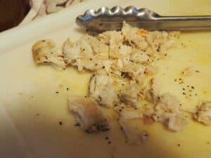 Cut up chicken breast on a white cutting board with silver tongs.