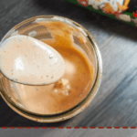 A jar of light orange Raising Cane's dipping sauce with a metal spoon and a jar of Tony Chachere's Creole Seasoning on a gray wooden background.