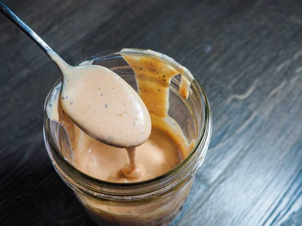 A jar of light orange Raising Cane's dipping sauce with a metal spoon on a gray wooden background.