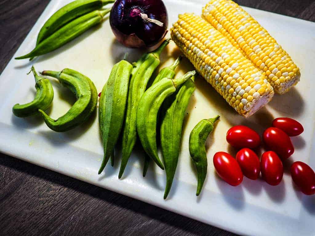 Corn, okra, cherry tomatoes, and a red onion with a white cutting board and knife on a gray background.