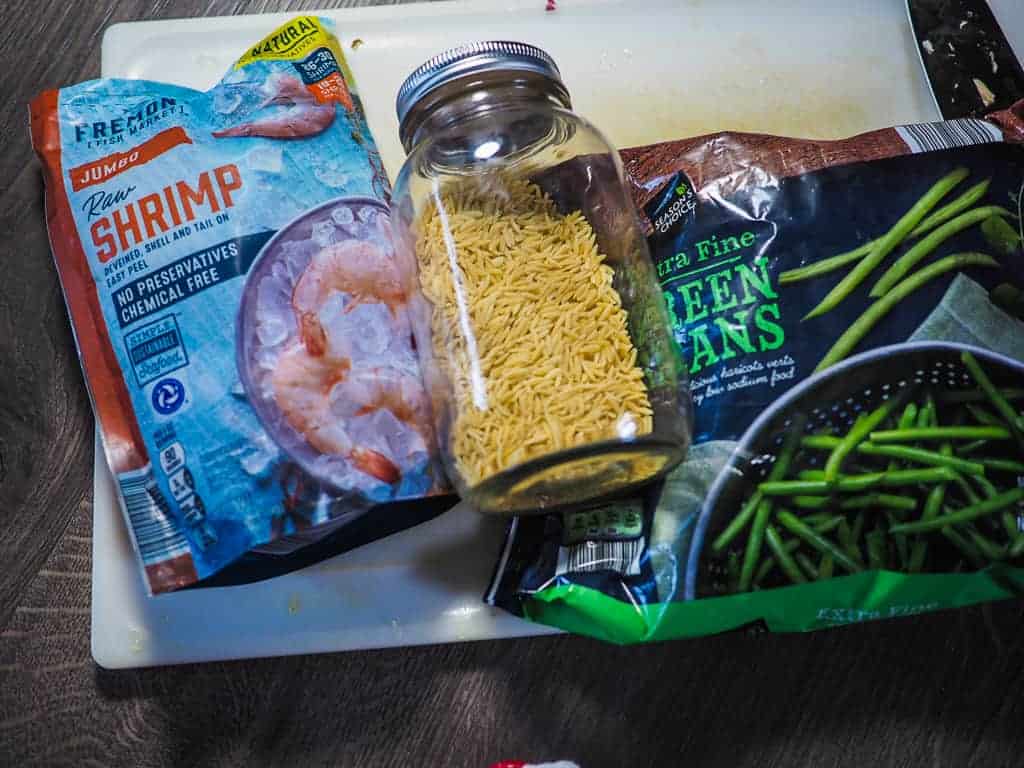 A bag of shrimp, a bag of green beans, and a jar of orzo on a gray background.