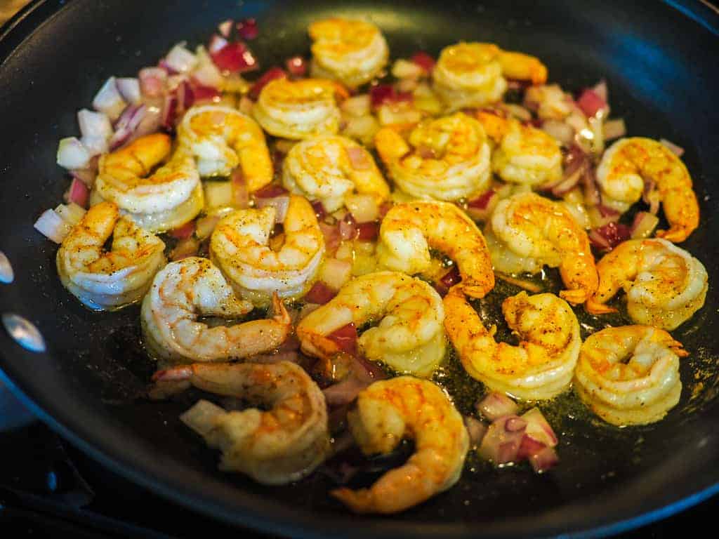 A saute pan with red onion and cooked shrimp in butter with a purple spatula.