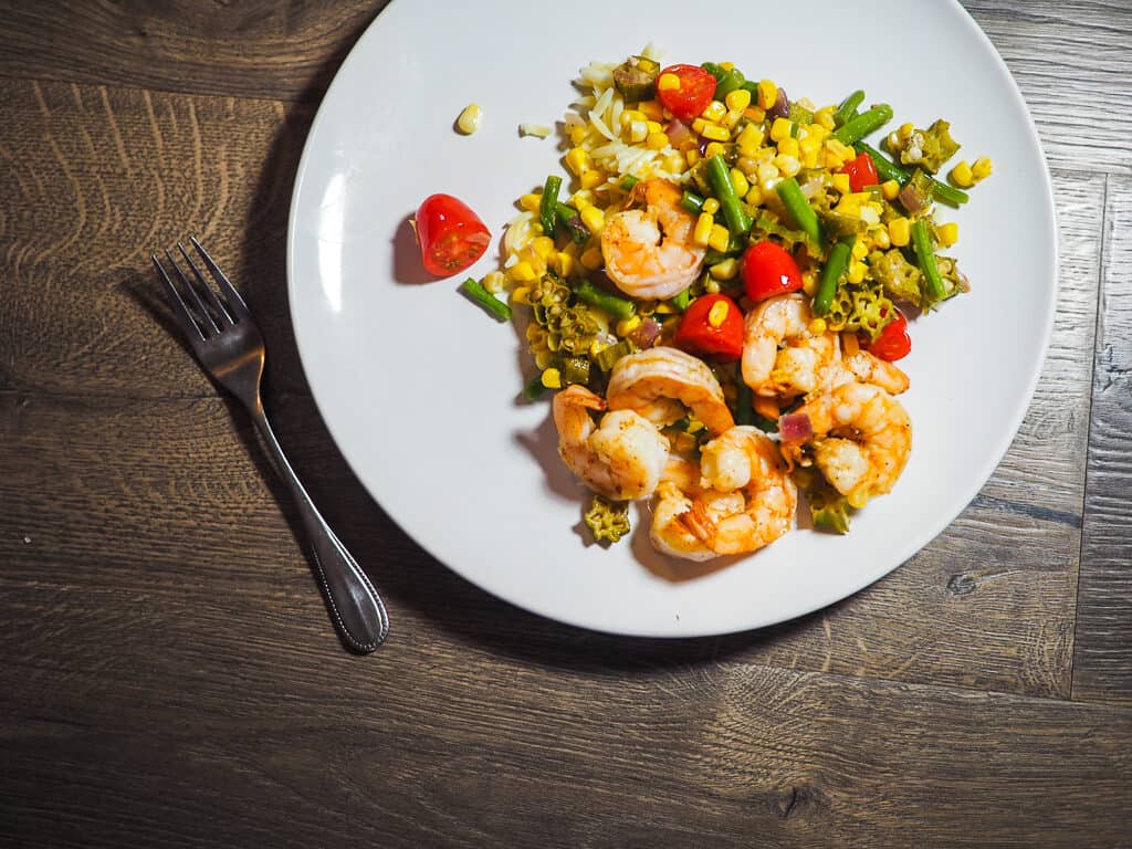 A white dish with shrimp, tomatoes, corn, and okra and a fork on a gray background.