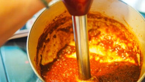 A white pot with reddish brown barbecue sauce simmering in it and an immersion blender pureeing it.