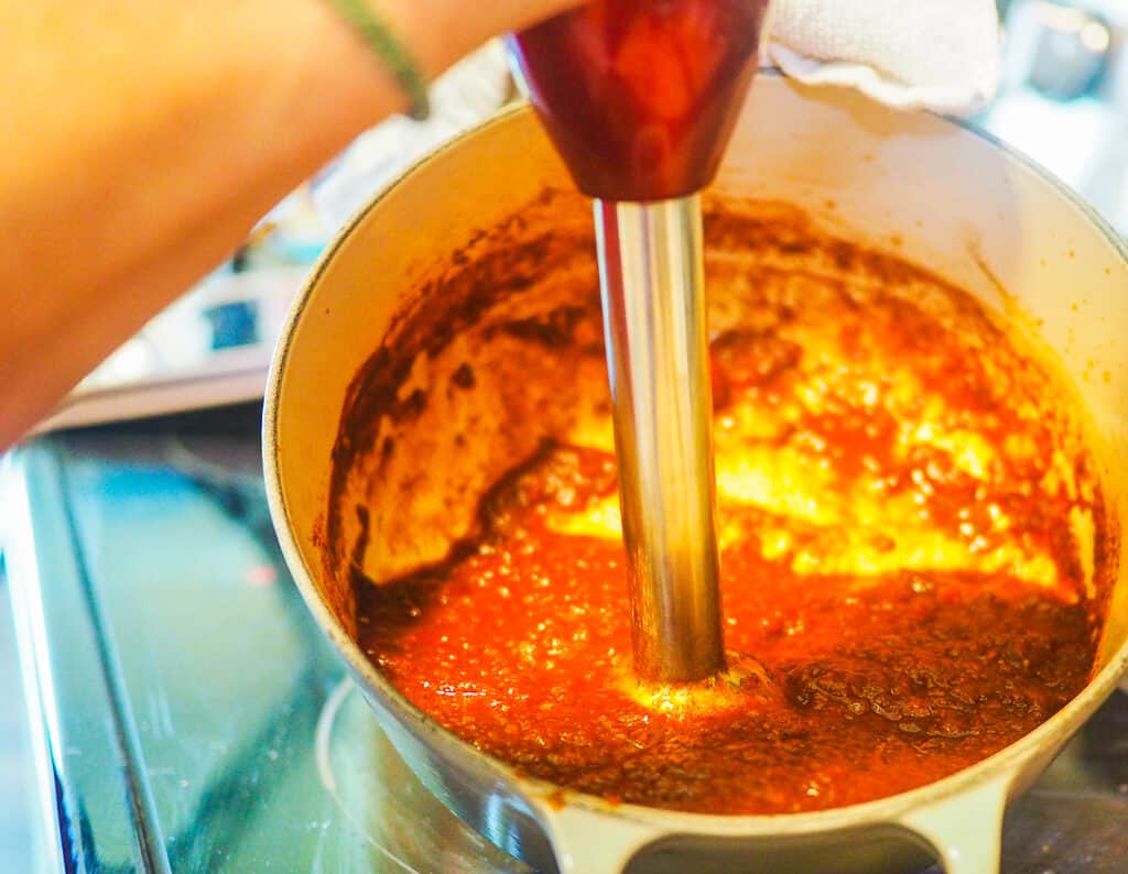 A white pot with reddish brown barbecue sauce simmering in it and an immersion blender pureeing it.