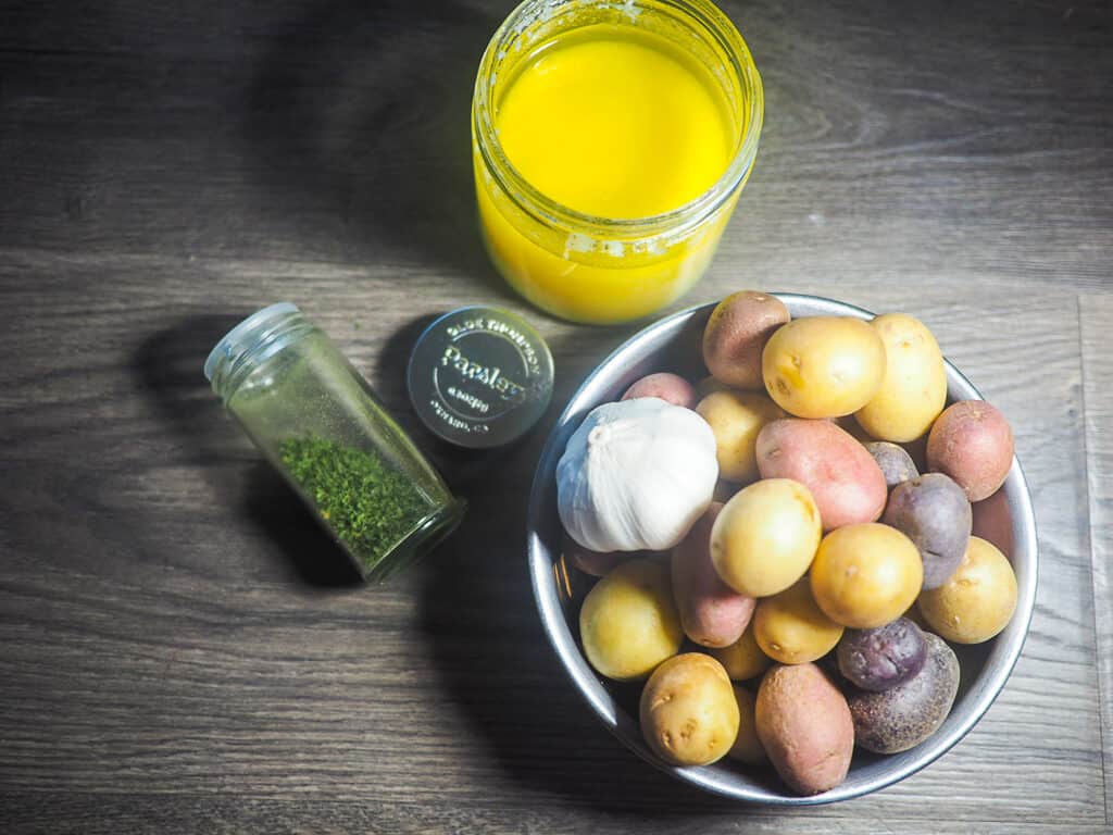 A jar of parsley, a jar of ghee, and a metal bowl of baby potatoes with a head of garlic on a gray wood background.