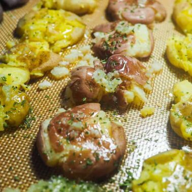 Smashed baby potatoes on a non-stick baking mat with ghee, garlic, and parsley.