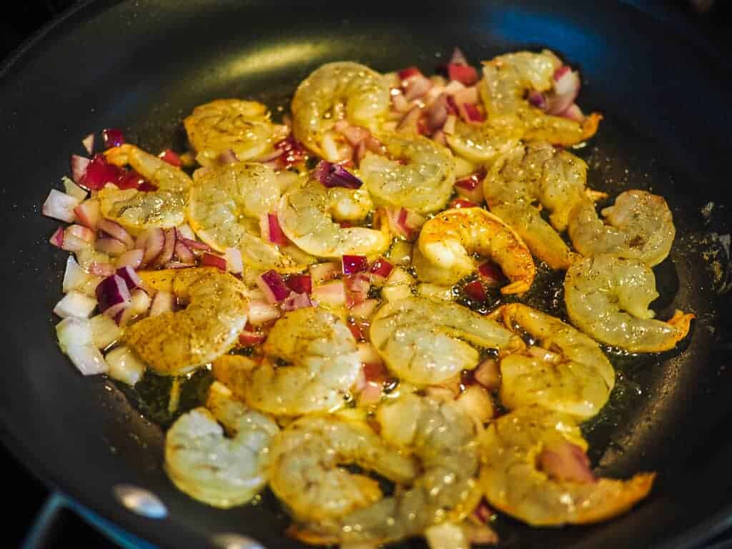A saute pan with red onion and shrimp in butter with a purple spatula.