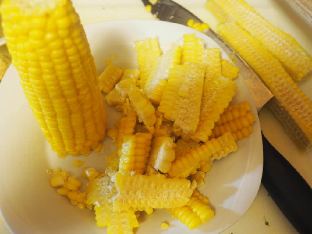 Corn kernels and a cob on a white cutting board with a knife.