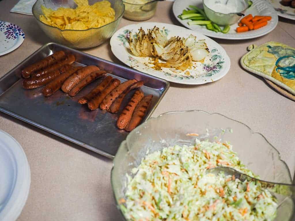 A kitchen counter with hot dogs, coleslaw, a bloomin onion, and dishes of vegetables.