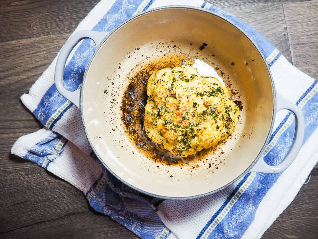 A roasted cauliflower in a gray dutch oven with a blue and white dish towel.