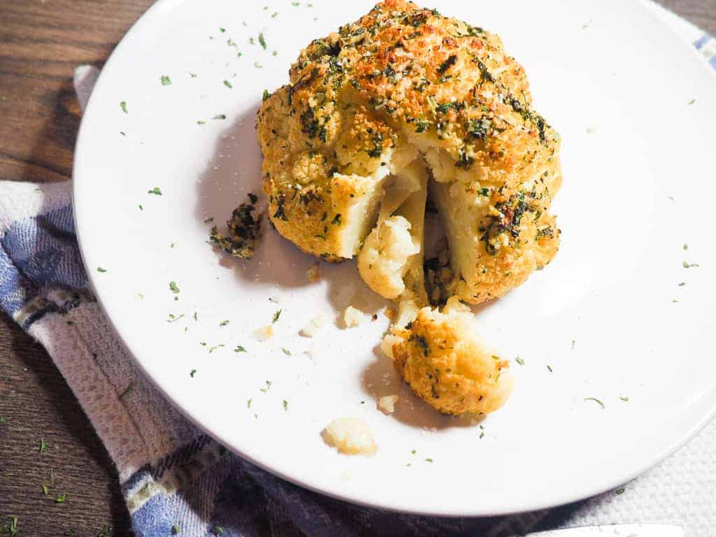 A whole head of roasted cauliflower on a white plate with a piece cut out on a blue and white dish towel.