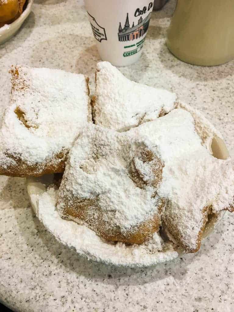 A plate of beignets covered in powdered sugar with a paper cup of cafe au lait from Cafe du Monde in New Orleans.