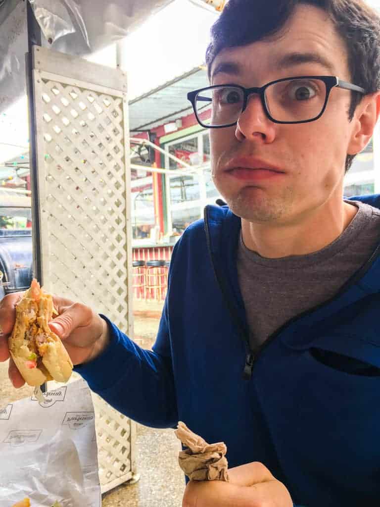 A man in a blue sweater, wearing glasses and making a funny face while eating a fried shrimp po-boy.