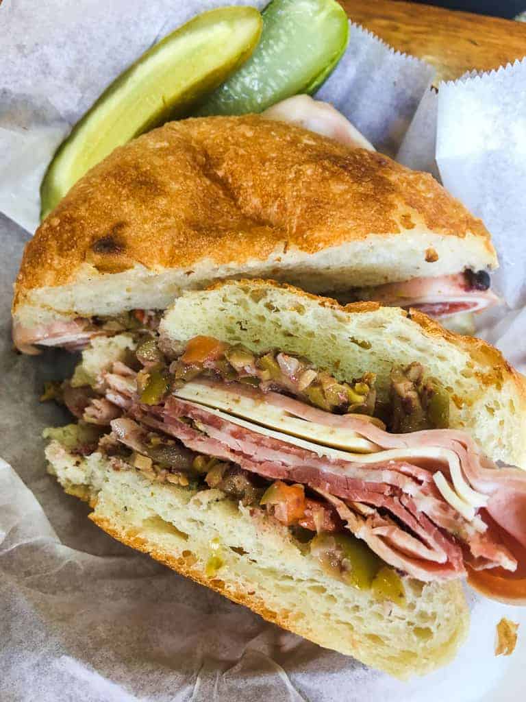 A muffaletta with meats and olive spread in a basket and a pickle from Stein's Deli in New Orleans.
