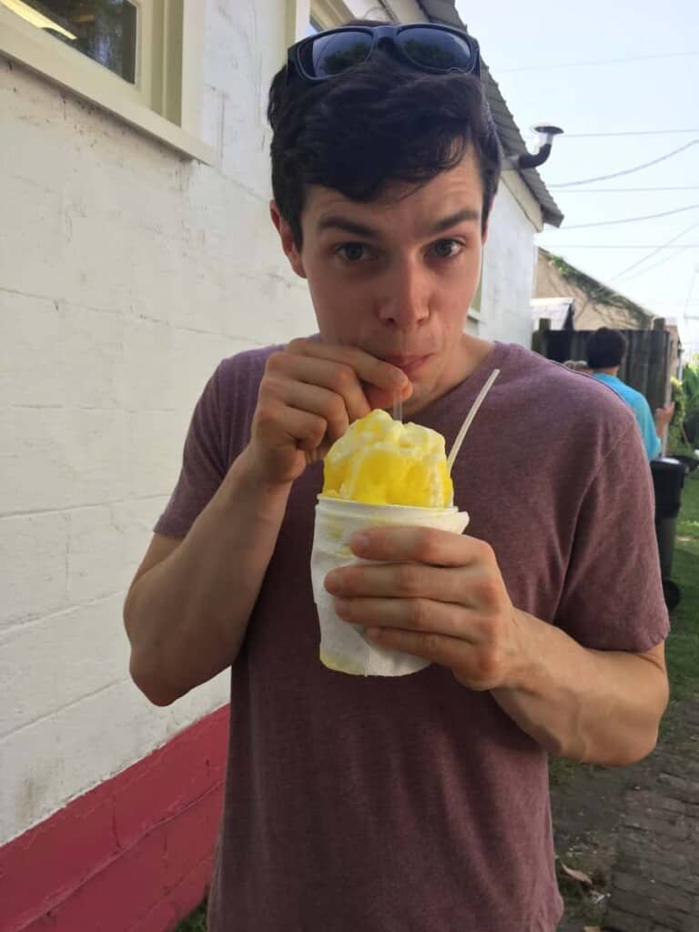 A brown haired man in a red shirt sipping a yellow snoball through a straw.
