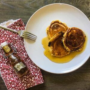 A white plate with three paleo pancakes soaked in maple syrup and a red napkin with a small bottle of maple syrup.