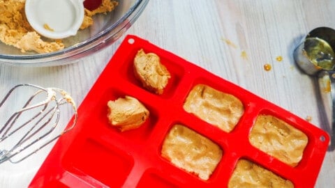A red silicone loaf baking sheet being filled with peanut butter perfect bars and a mixing bowl with a red spatula and a measuring cup in it.