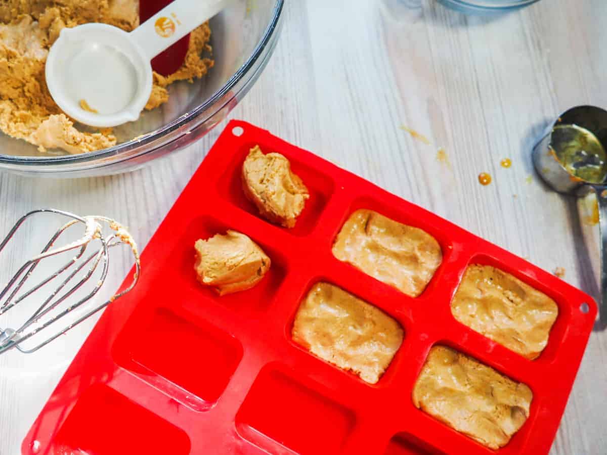 A red silicone loaf baking sheet being filled with peanut butter perfect bars and a mixing bowl with a red spatula and a measuring cup in it.