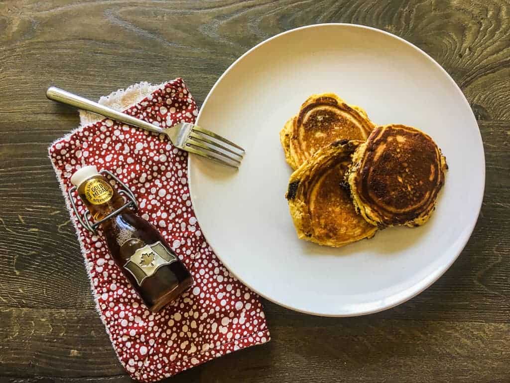A white plate with three paleo pancakes and a red napkin with a small bottle of maple syrup.