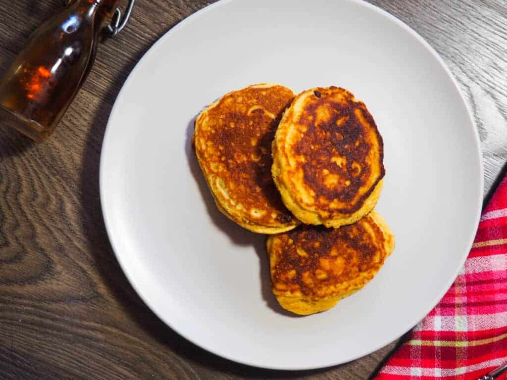 Three sweet potato paleo pancakes on a white plate with a red checkered napkin and small bottle of maple syrup.