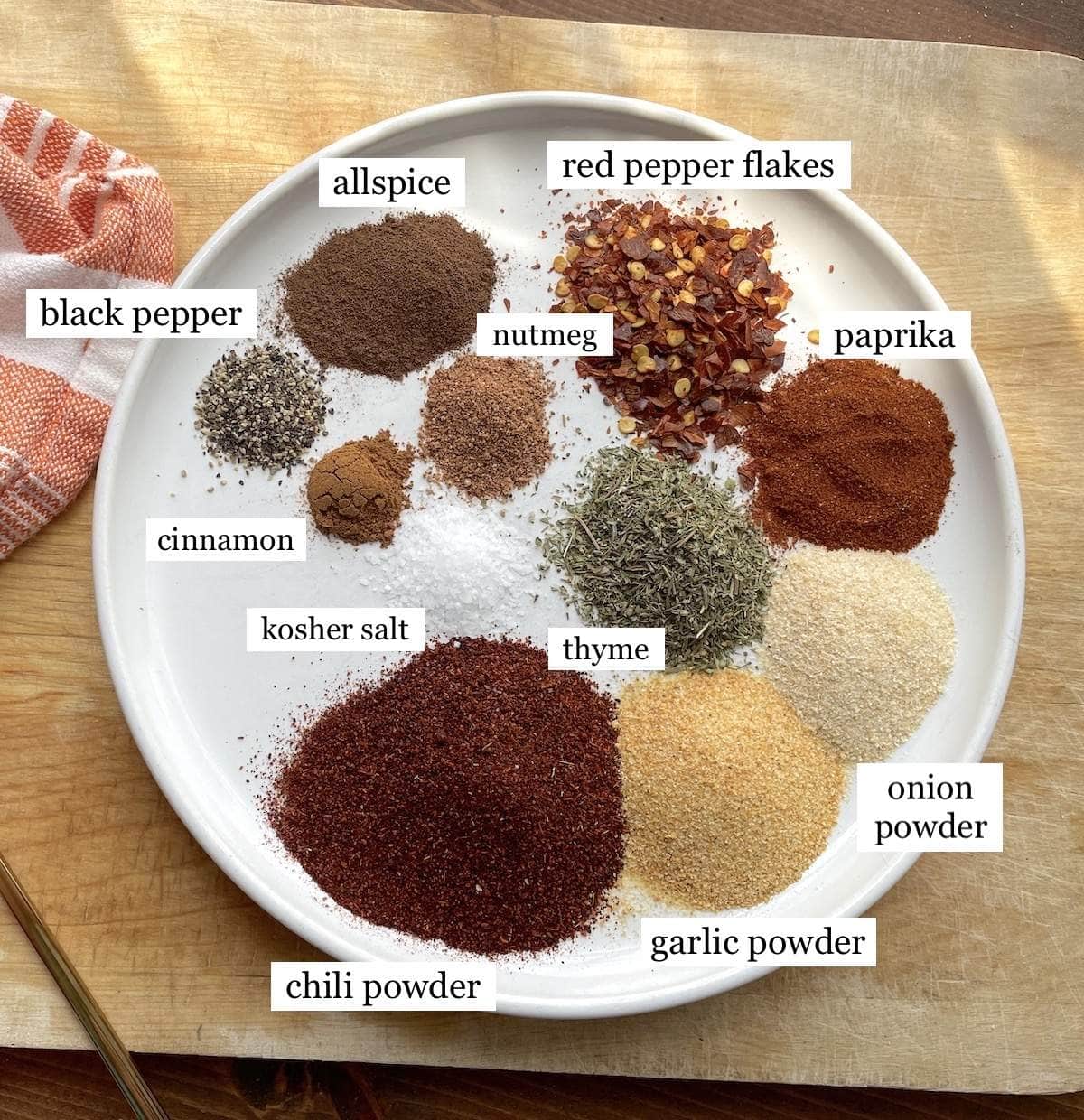 The ingredients of Jamaican Jerk Seasoning laid out in small colorful bowls and labeled on a marble backdrop with a blue dish towel.