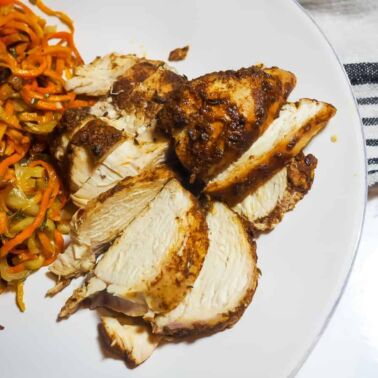 A left aligned white plate with crispy sweet potato noodles and sliced Jamaican Jerk Chicken with a gray and navy dish towel.