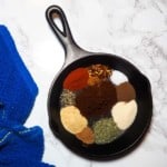 A small black cast iron pan with the unmixed ingredients for making Jamaican Jerk Seasoning and a blue dish cloth.