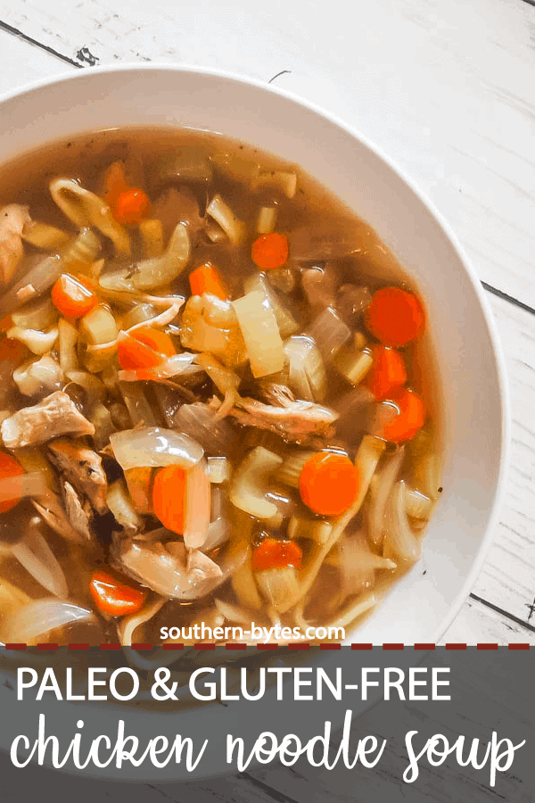 A pin image of a white bowl of paleo chicken noodle soup with carrots, celery, and onions on a white wooden background.