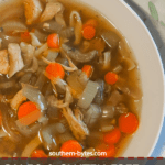 A pin image of a white bowl of paleo chicken noodle soup with carrots, celery, and onions on a white wooden background with a blue and white tea towel.