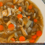 A pin image of a white bowl of paleo chicken noodle soup with carrots, celery, and onions on a white wooden background with a blue and white tea towel.
