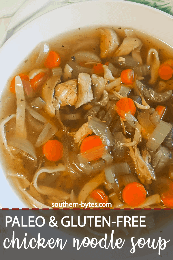 A pin image of a white bowl of paleo chicken noodle soup with carrots, celery, and onions on a white wooden background with a blue and white tea towel.