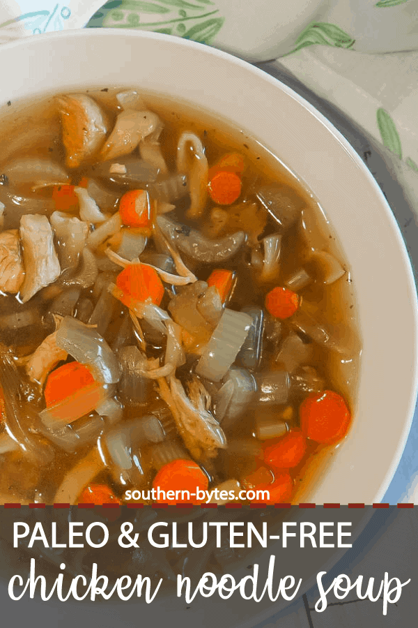 A pin image of a white bowl of paleo chicken noodle soup with carrots, celery, and onions on a white wooden background with a blue and white tea towel.