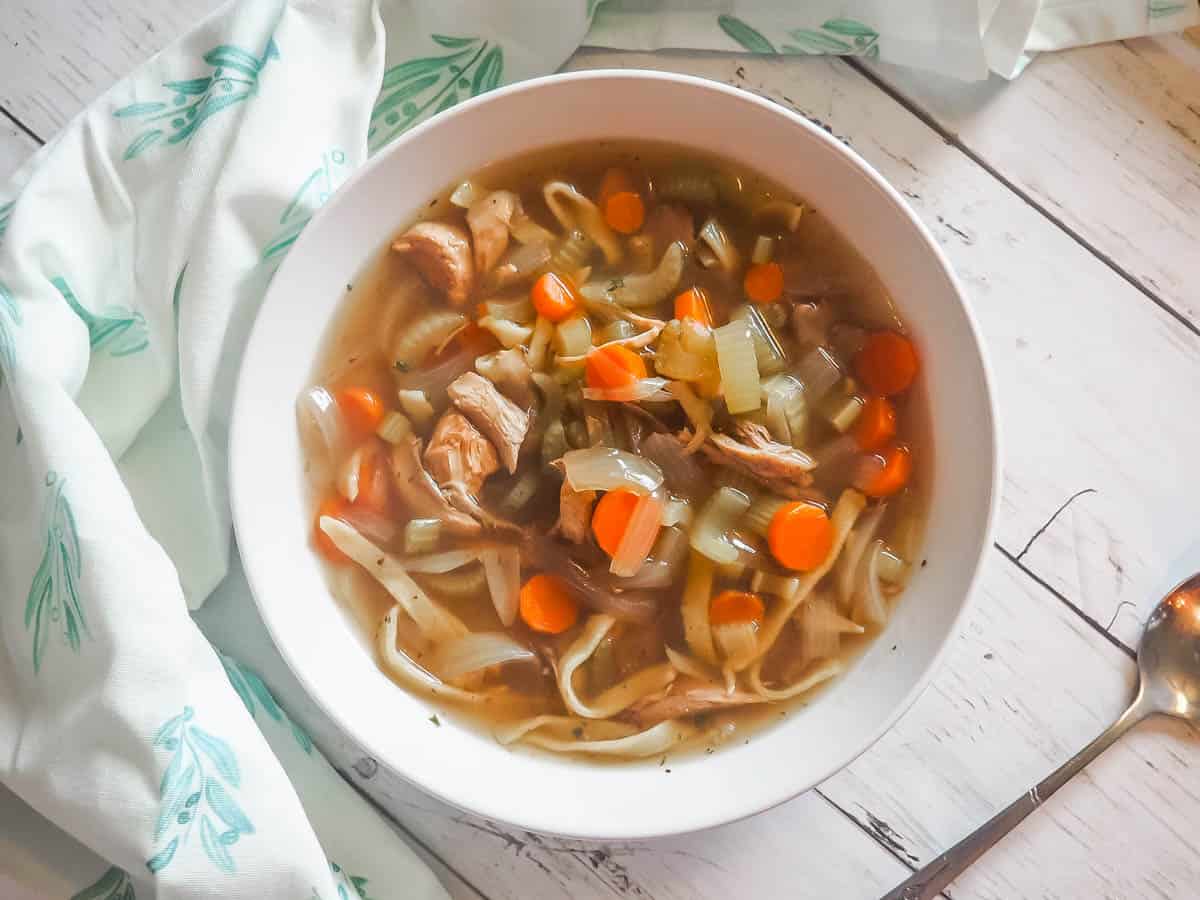 A white bowl of paleo chicken noodle soup with carrots, celery, and onions on a white wooden background with a spoon and blue and white tea towel.