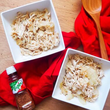 Two white bowls of shredded chicken and onions with chili lime seasoning with a red cloth napkin and a bottle of Tajin seasoning on a wooden background.