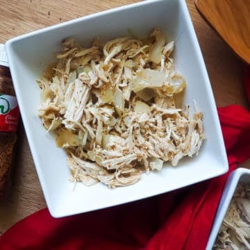 A white bowl of diced chicken and onions with chili lime seasoning with a red cloth napkin and a bottle of Tajin seasoning on a gray wooden background.