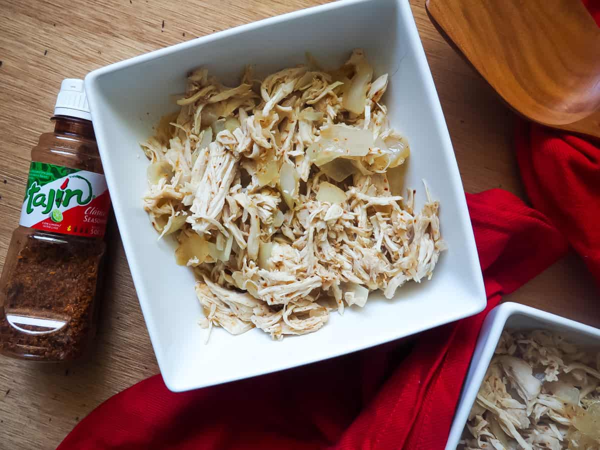 A white bowl of diced chicken and onions with chili lime seasoning with a red cloth napkin and a bottle of Tajin seasoning on a gray wooden background.