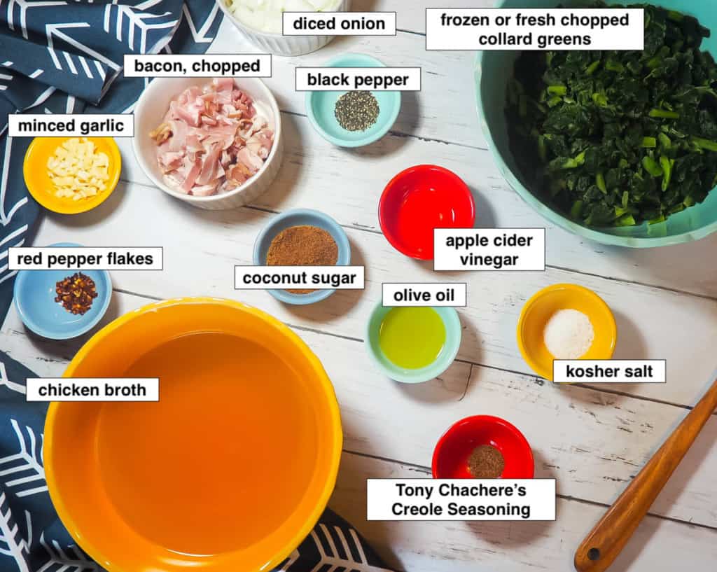 The ingredients of collard greens laid out and labeled on a white wood background: bacon, collard greens, chicken broth, red pepper flakes, coconut sugar, apple cider vinegar, kosher salt, olive oil, black pepper, diced onion, and minced garlic.