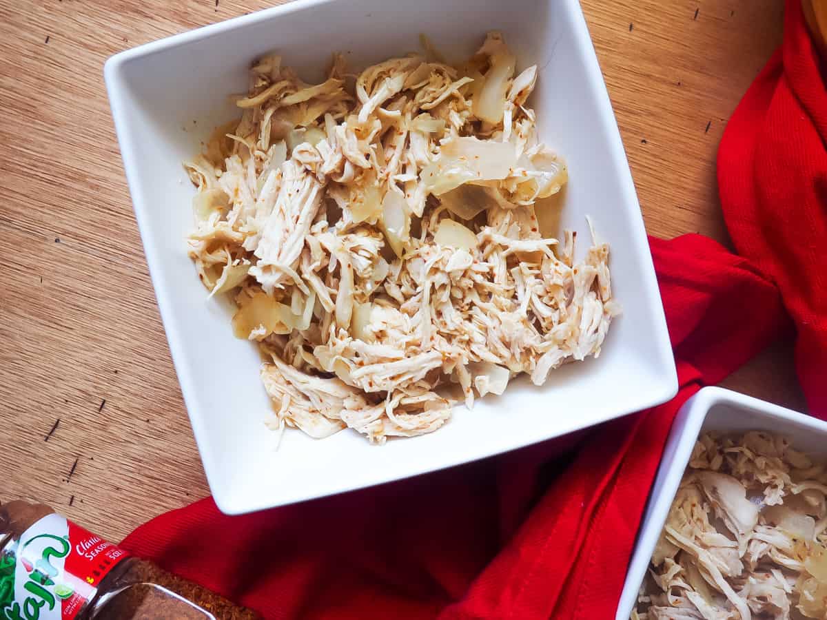 A white bowl of diced chicken and onions with chili lime seasoning with a red cloth napkin and a bottle of Tajin seasoning on a gray wooden background.