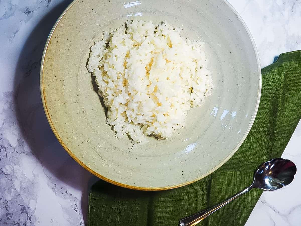 A bowl of white rice on a marble background with a green napkin and a spoon.