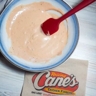 A pin image blue and white bowl of orange Raising Cane's dipping sauce with a red spatula and a Cane's napkin.