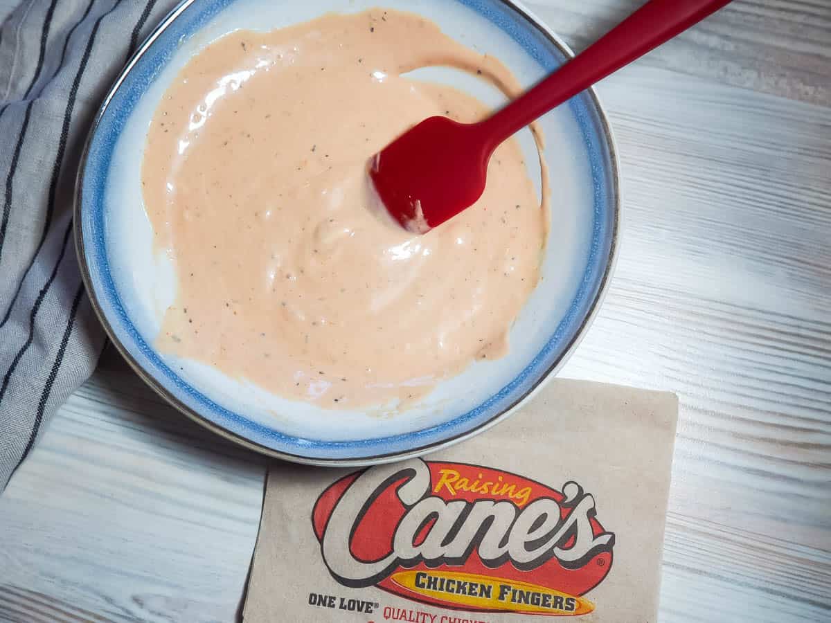 A pin image blue and white bowl of orange Raising Cane's dipping sauce with a red spatula and a Cane's napkin.