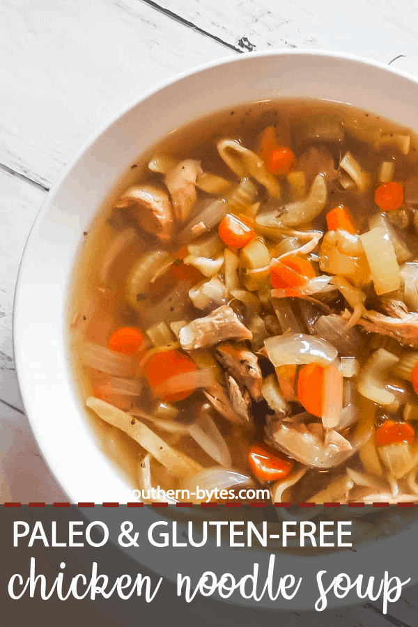 A pin image of a white bowl of paleo chicken noodle soup with carrots, celery, and onions on a white wooden background.