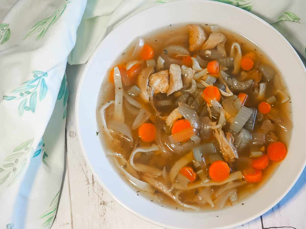 A white bowl of paleo chicken noodle soup with carrots, celery, and onions on a white wooden background with a blue and white tea towel.