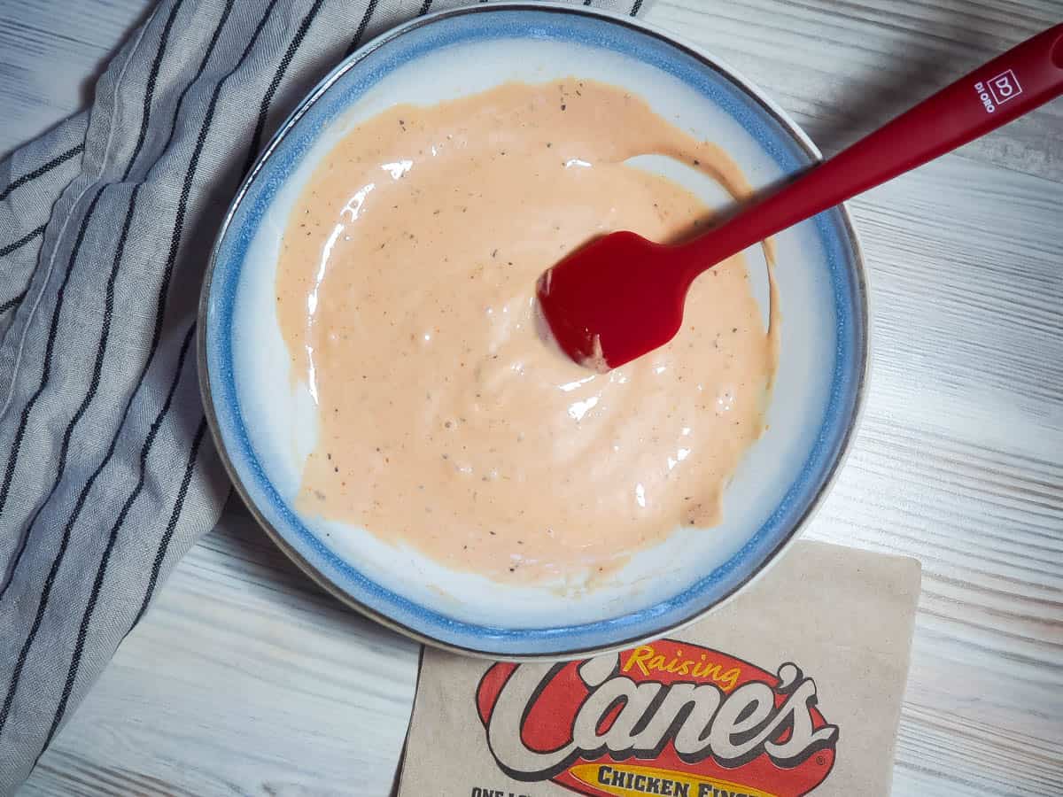 A blue and white bowl of orange Raising Cane's dipping sauce with a red spatula and a Cane's napkin.