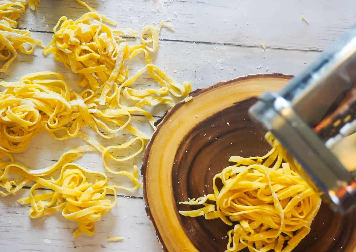 Piles of paleo pasta and a pile of pasta coming out of a pasta maker on a wood cutting board.