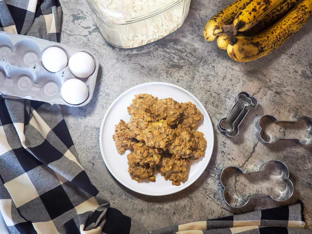 A plate of peanut butter and banana dog treats with dog treat cookie cutters, eggs, oats, and bananas.