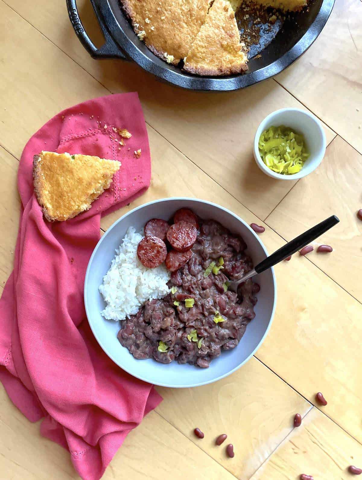 A bowl of red beans and rice with sliced andouille, a small bowl of pepperoncini peppers, a slice of cornbread, and a pan of cornbread.